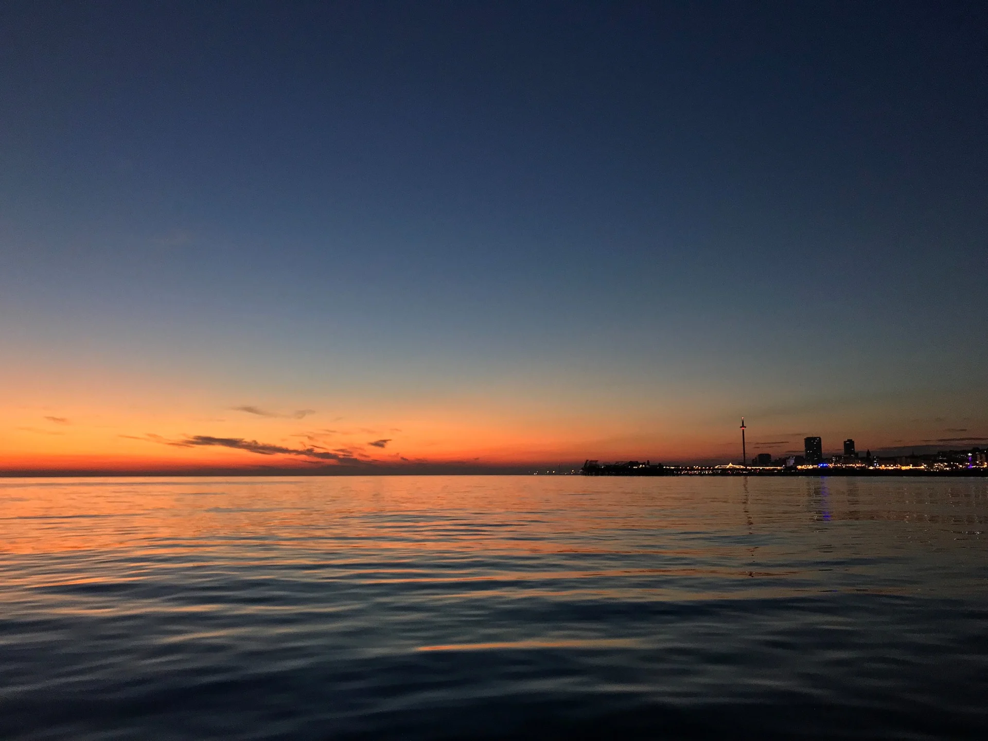 Brighton seafront at sunset, with calm water, glowing horizon light, and the distant city skyline.